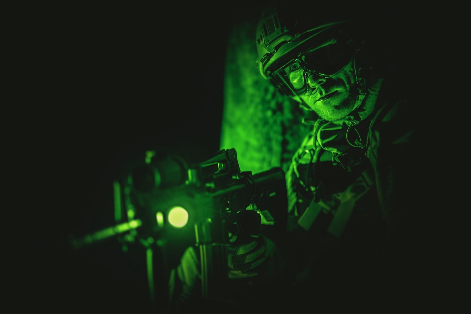Night-vision view of an operator in a helmet aiming a rifle with an illuminated optic